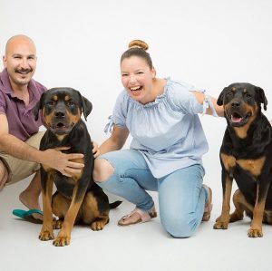 A man and woman smiling with their arms around two Rottweilers.