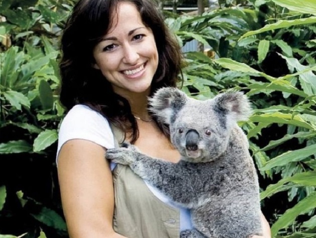 A brunette woman holding a koala in front of some trees.